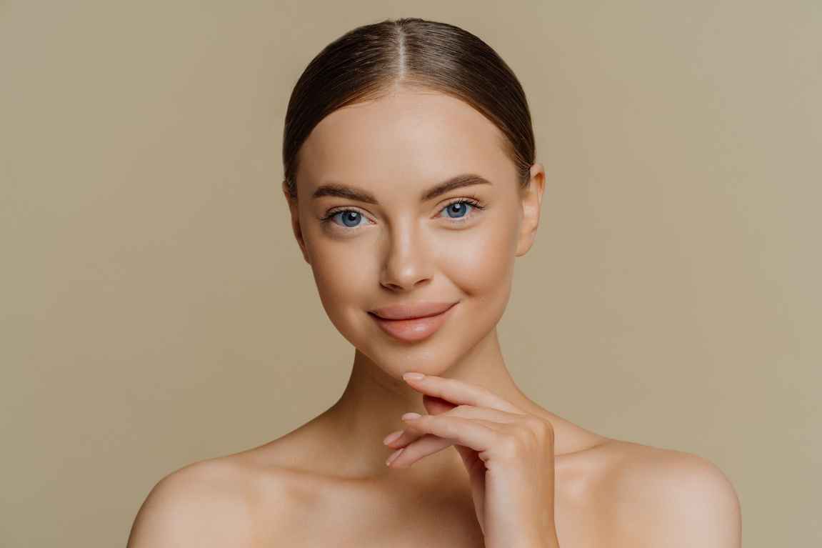 Headshot of Beautiful Brunette Woman Touches Jawline Looks Directly at Camera Stands Shirtless Has Natural Beauty Isolated over Brown Background. People Wellness Cosmetology Skin Care Concept